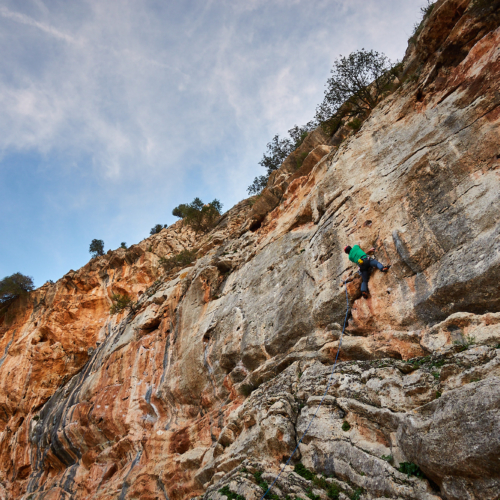 A climber sport climbing on a grey and orange limestone cliff in southern Spain with blue sky and thin clouds above
