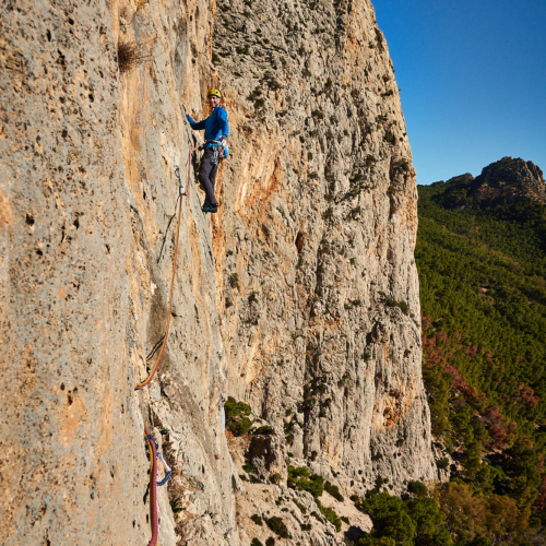 A climber sport climbing on the fifth pitch of a route called Amptrax on a limestone cliff in southern Spain with blue sky above and green trees below