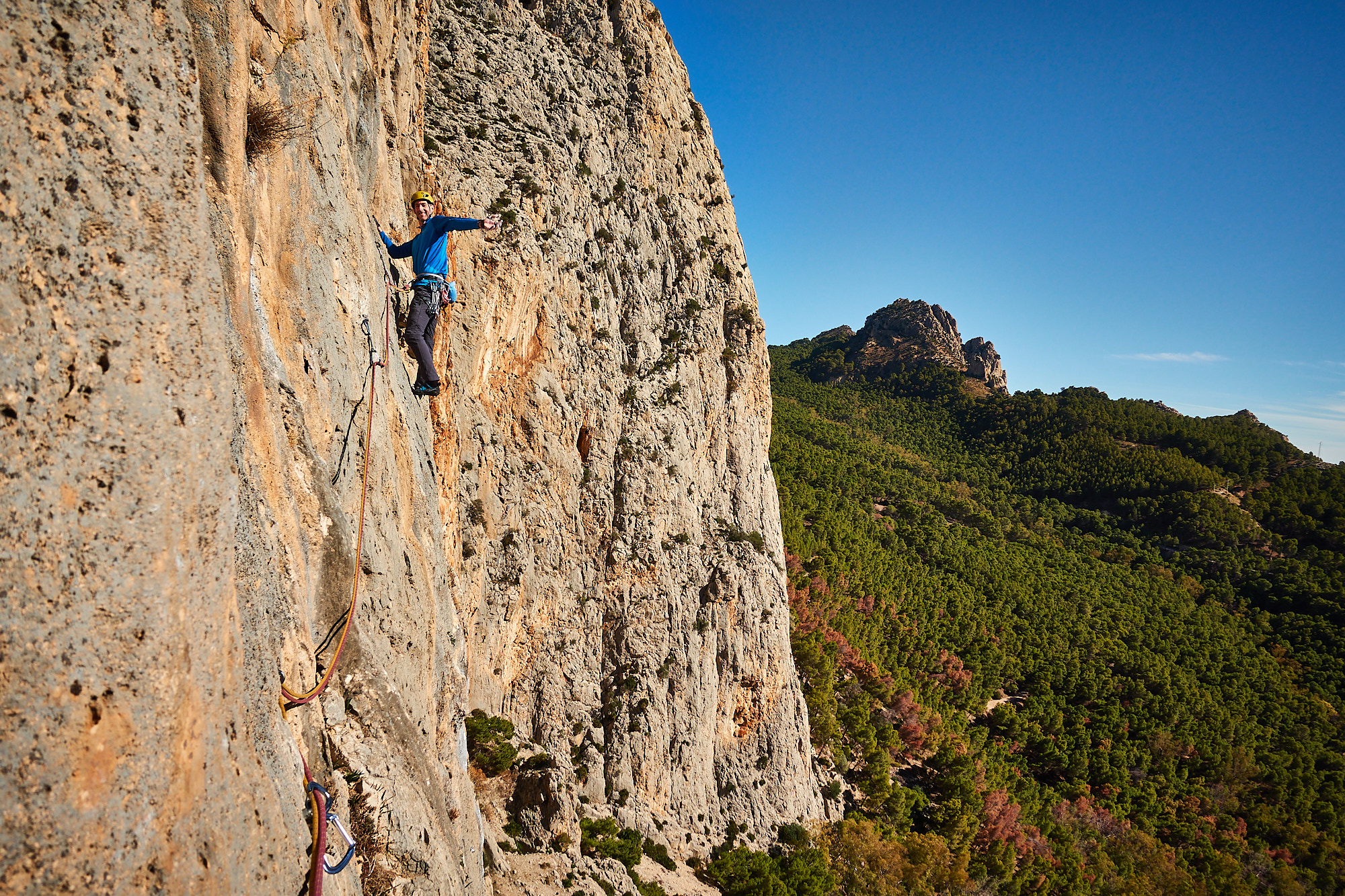 A climber sport climbing on the fifth pitch of a route called Amptrax on a limestone cliff in southern Spain with blue sky above and green trees below