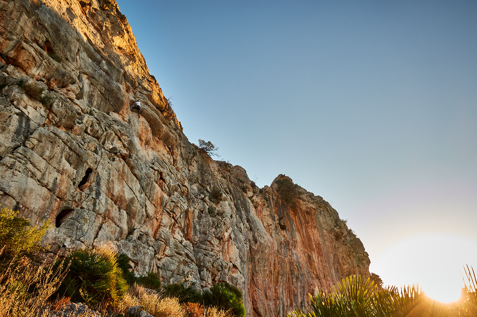 A climber sport climbing on a grey and orange limestone cliff in southern Spain with blue sky above and green hill and a lake behind
