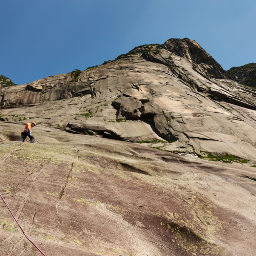 A climber in orange on a steep brown and pink granite slab with blue sky above