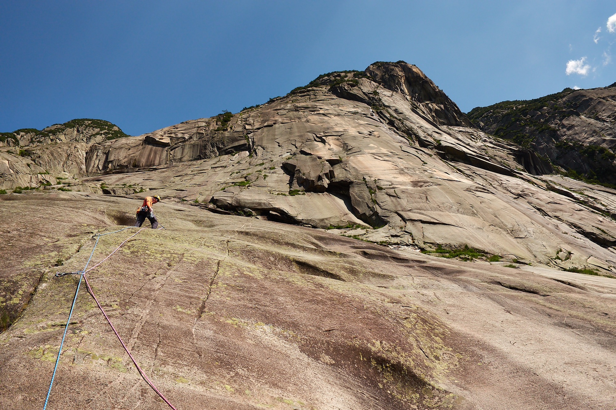 A climber in orange on a steep brown and pink granite slab with blue sky above