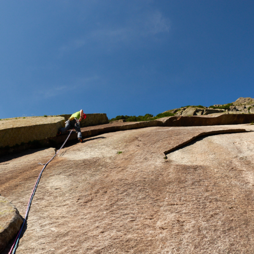 A climber in green in a corner on the left side of smooth brown and white granite slab with an overlap above