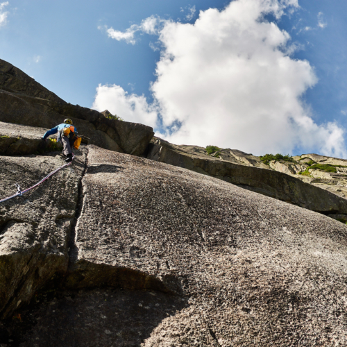 A climber in blue climbing a crack on the left of a smooth grey granite slab with overlaps above