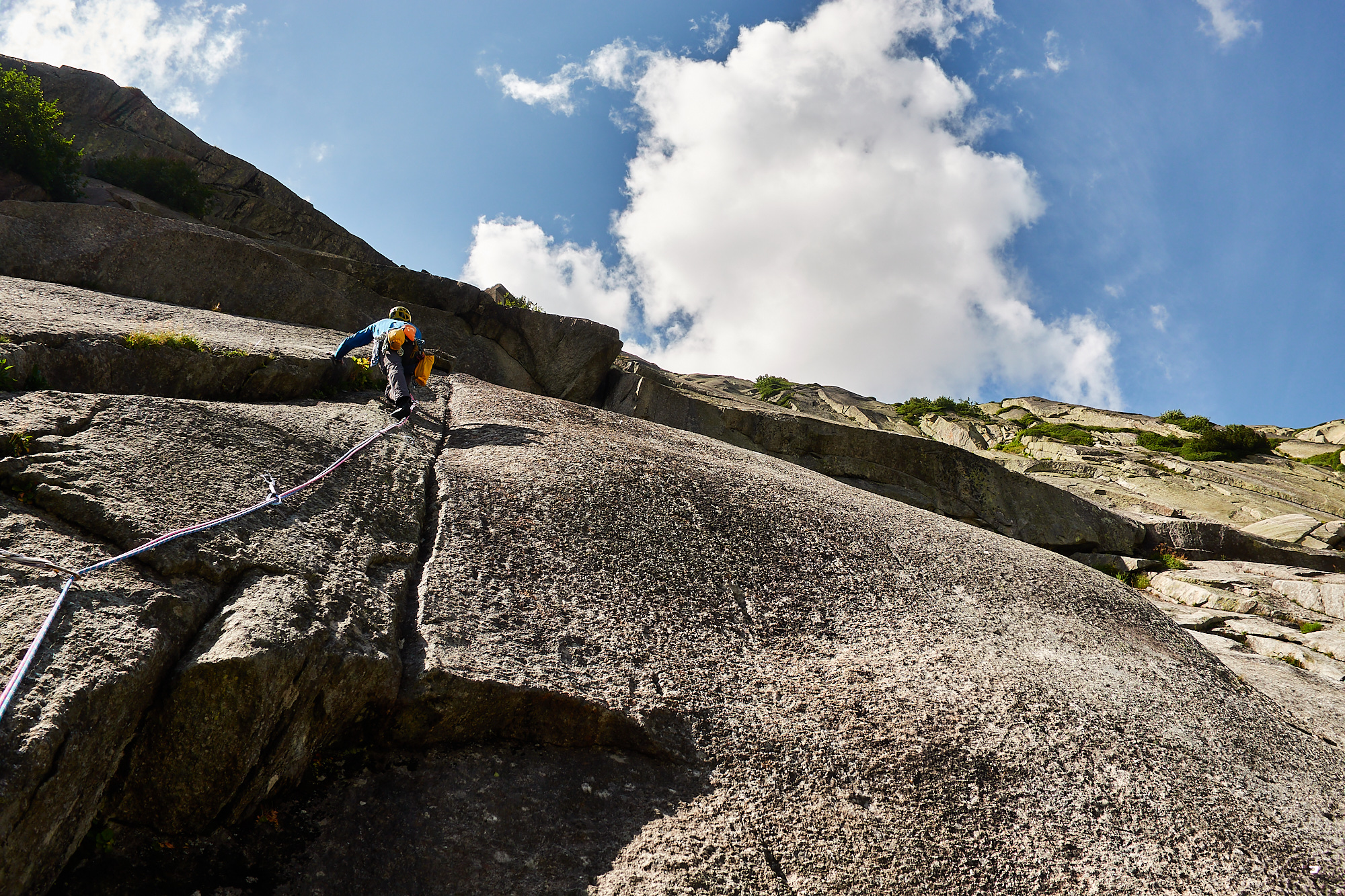 A climber in blue climbing a crack on the left of a smooth grey granite slab with overlaps above