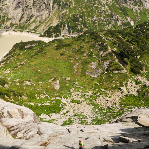 A view down a steep white granite slab of a climber abseiling down with alpine meadows and lakes below