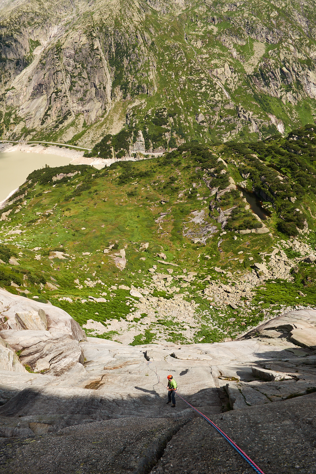 A view down a steep white granite slab of a climber abseiling down with alpine meadows and lakes below