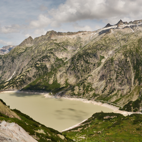 A view from a mountainside looking over an artificial dammed lake towards granite cliffs and jagged mountain tops with clouds above