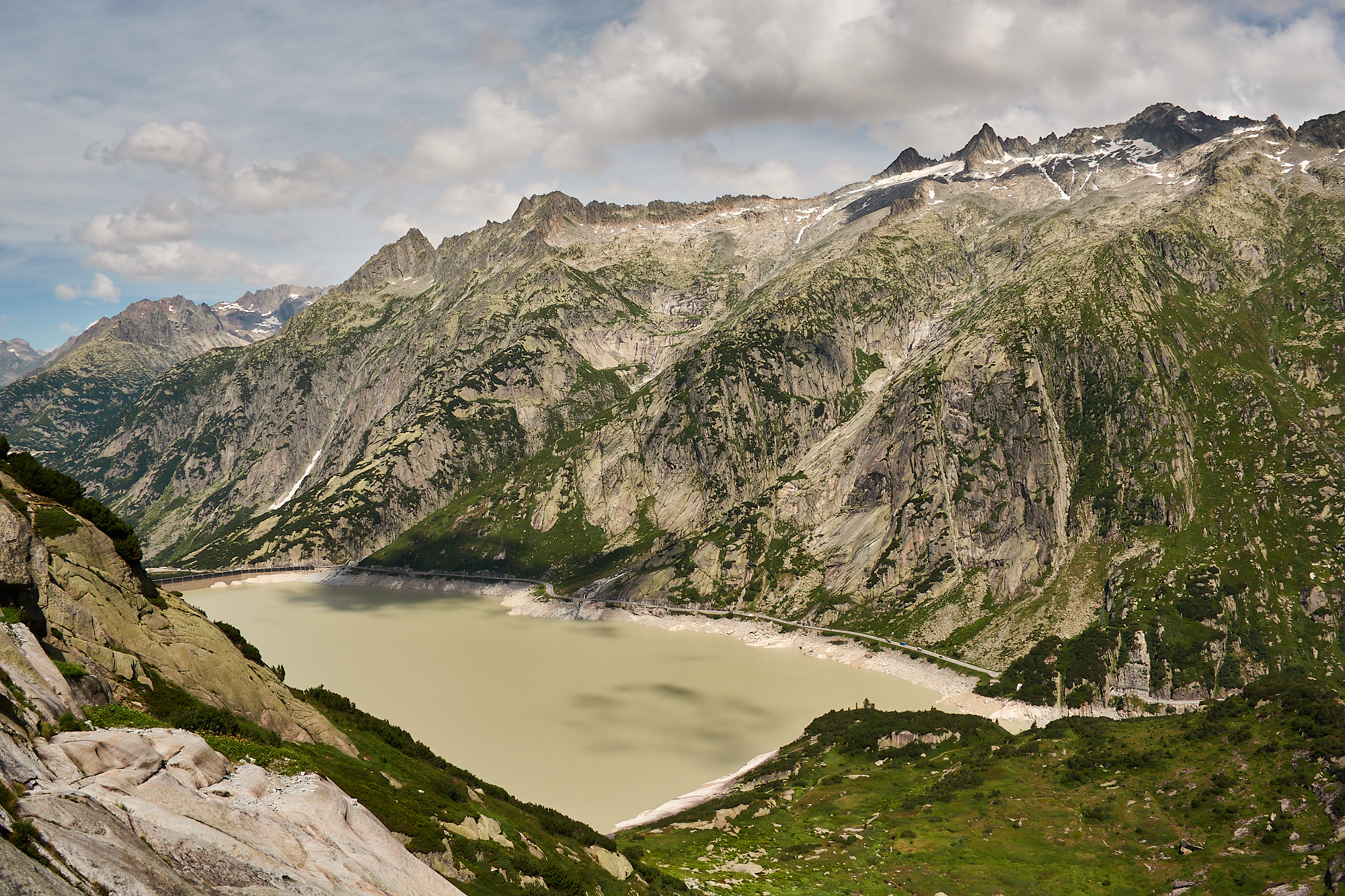 A view from a mountainside looking over an artificial dammed lake towards granite cliffs and jagged mountain tops with clouds above