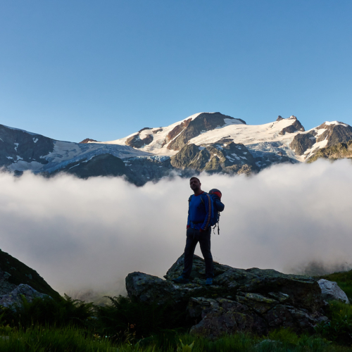 A mountaineer standing on a boulder above a cloud inversion in the valley with alpine peaks covered in glaciers and blue sky above