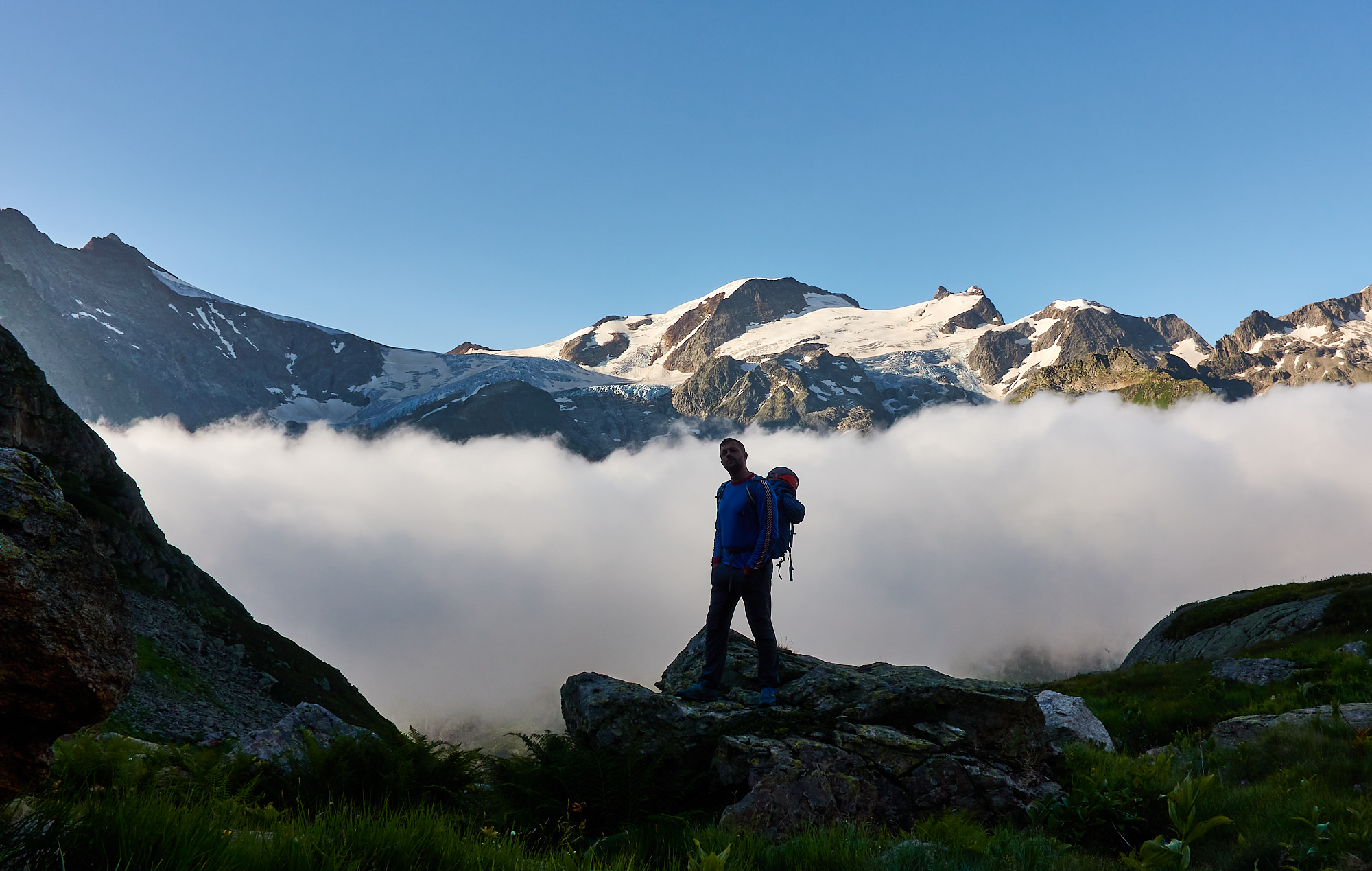 A mountaineer standing on a boulder above a cloud inversion in the valley with alpine peaks covered in glaciers and blue sky above