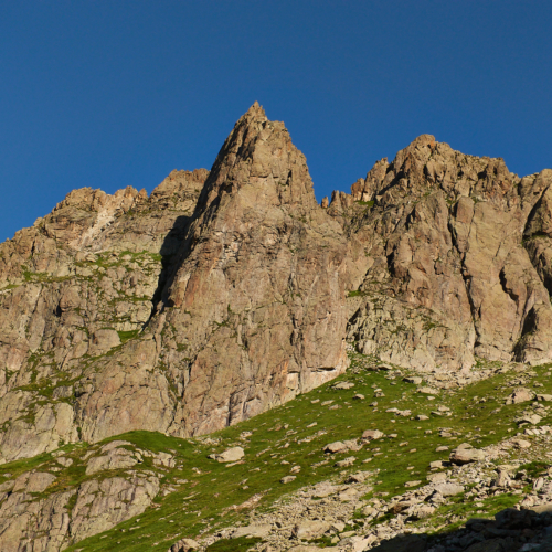 A view of a jagged, pyramid-shaped, white and brown rock peak above a green alpine meadow, below a blue sky