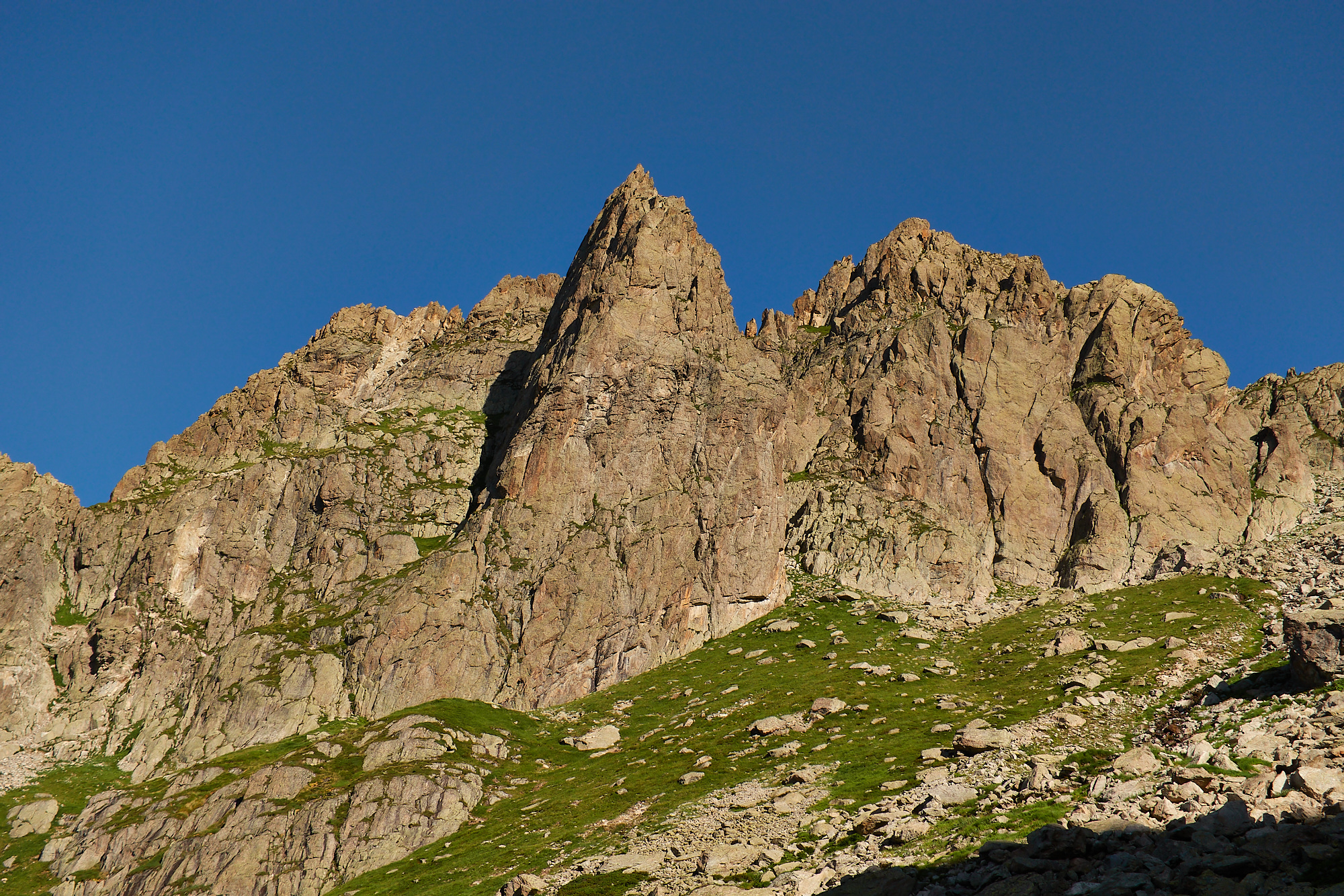 A view of a jagged, pyramid-shaped, white and brown rock peak above a green alpine meadow, below a blue sky