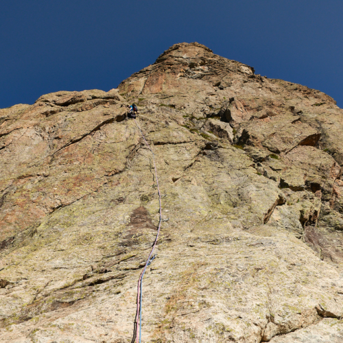 A climber in blue climbing a corner-crack on the left of a smooth white and brown slab with a steep rock face above