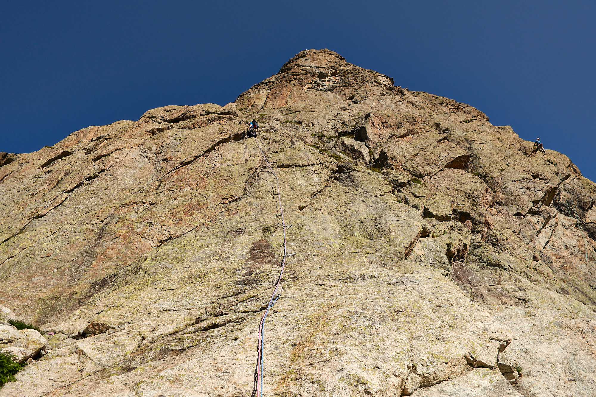 A climber in blue climbing a corner-crack on the left of a smooth white and brown slab with a steep rock face above