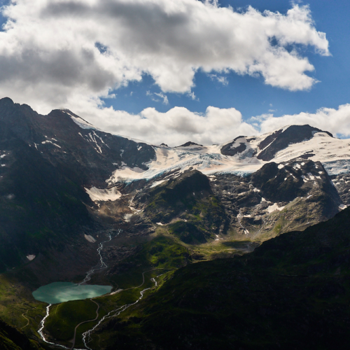 A dramatic view of alpine mountains with rocky summits and glaciated slopes above green alpine meadows and a green glacial lake with blue sky and clouds above