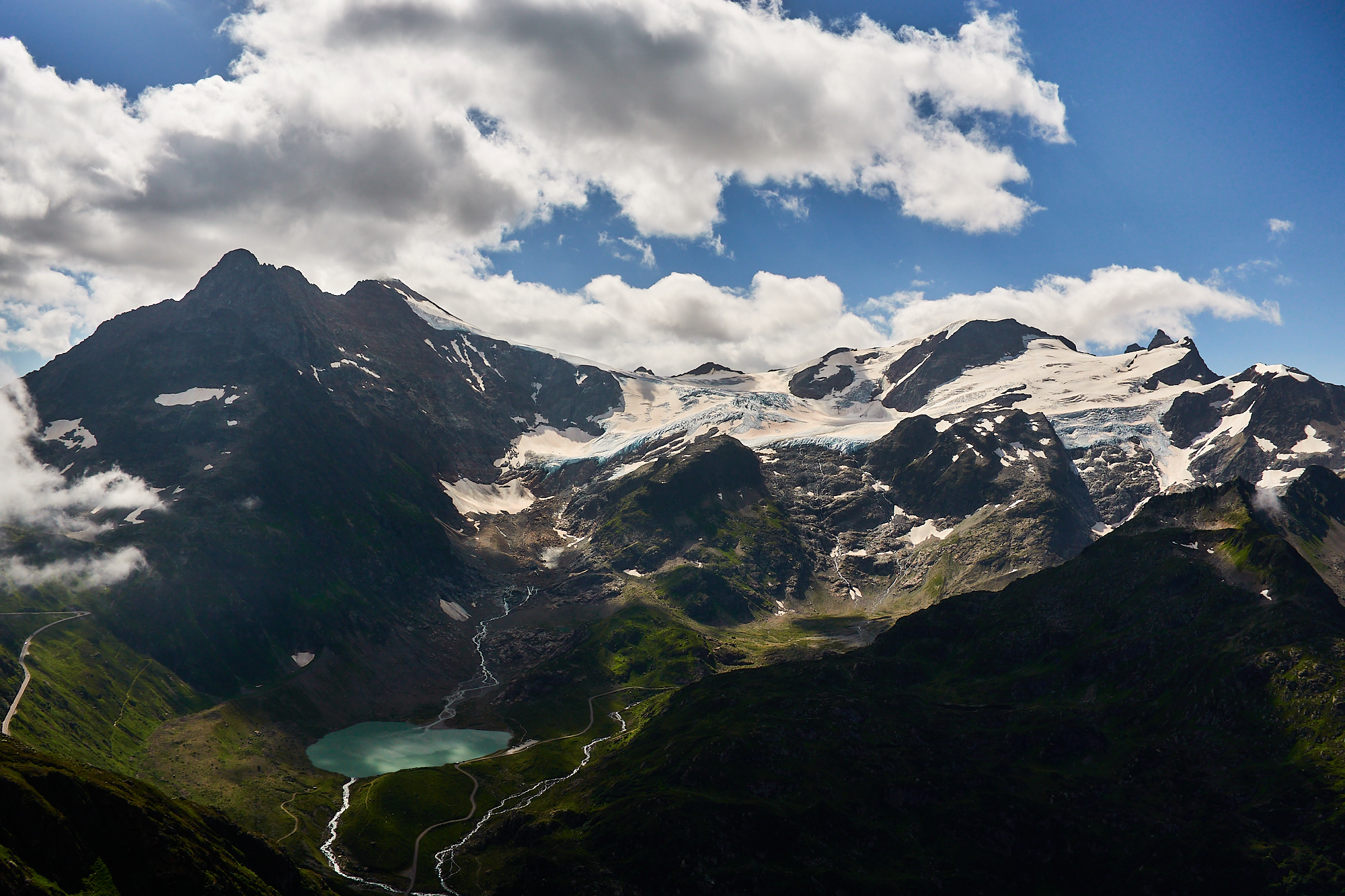 A dramatic view of alpine mountains with rocky summits and glaciated slopes above green alpine meadows and a green glacial lake with blue sky and clouds above