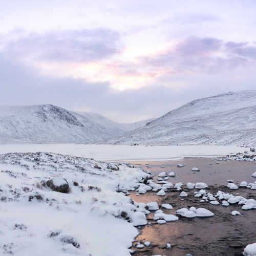 A wintery scottish mountain landscape of rounded hills, craggy cliffs and frozen lochs with a covering of snow