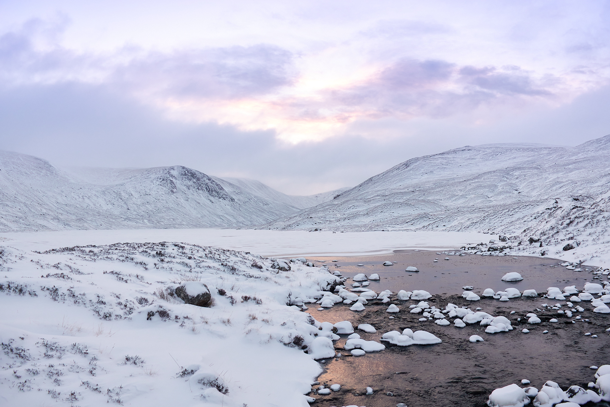 A wintery scottish mountain landscape of rounded hills, craggy cliffs and frozen lochs with a covering of snow