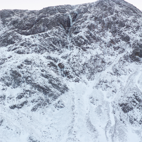 A wintery mountain rock face with snow patches below and a frozen lochan, split by gully containing steep blue icefalls.