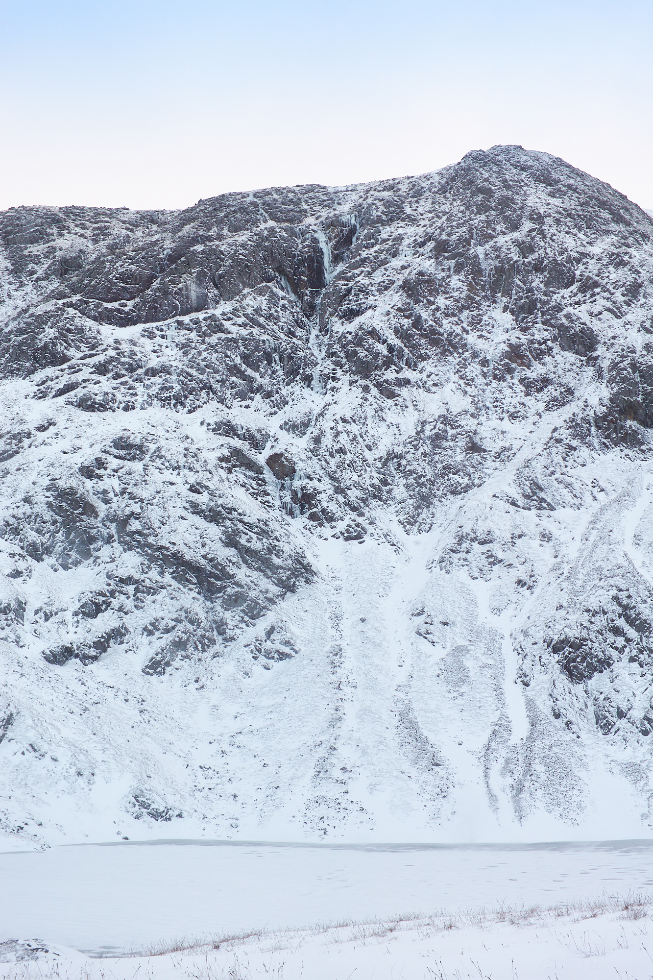 A wintery mountain rock face with snow patches below and a frozen lochan, split by gully containing steep blue icefalls.