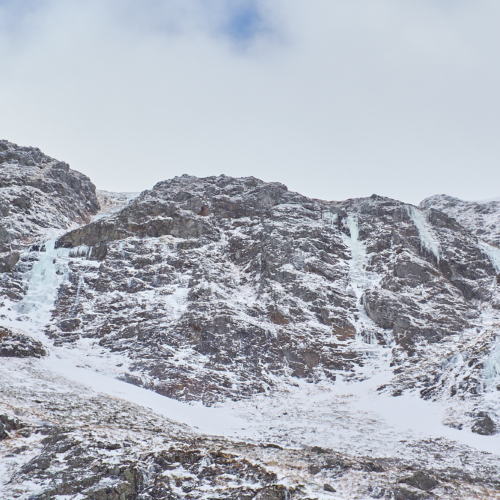 A wintery mountain rock face with snow patches below, split by three steep blue icefalls