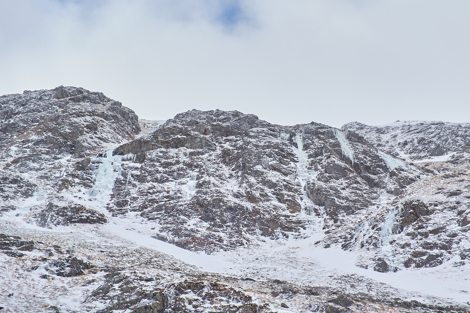 A wintery mountain rock face with snow patches below, split by three steep blue icefalls