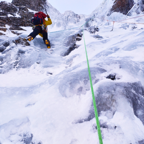 A climber in bright clothing swinging their ice axe as they climb a pitch of ice