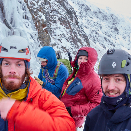 A group of climbers in brightly coloured winter clothing relaxing on a ledge at the base of a steep blue icefall