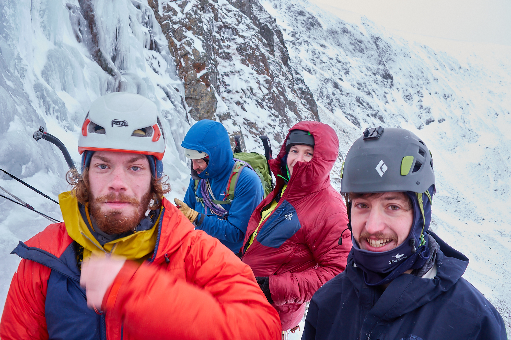 A group of climbers in brightly coloured winter clothing relaxing on a ledge at the base of a steep blue icefall