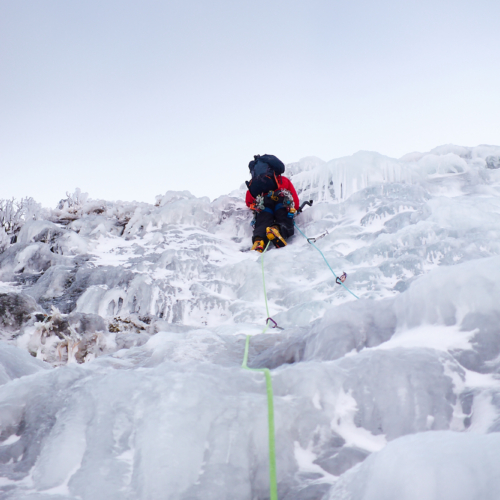 A climber in bright clothing swinging their ice axe as they climb a pitch of featured ice