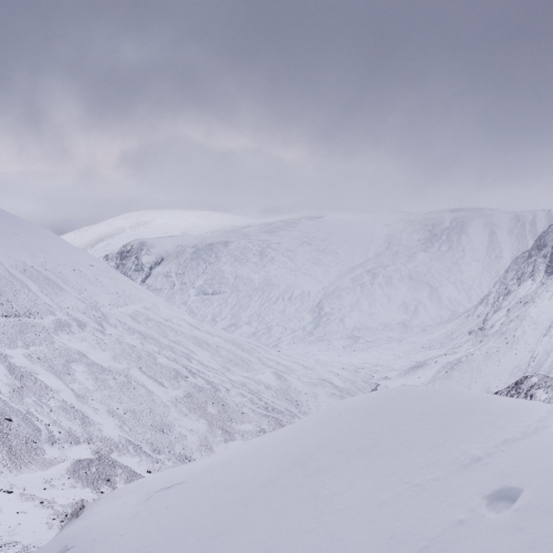 A snowy mountain landscape of rounded hills and dark craggy cliffs with ominous grey clouds above