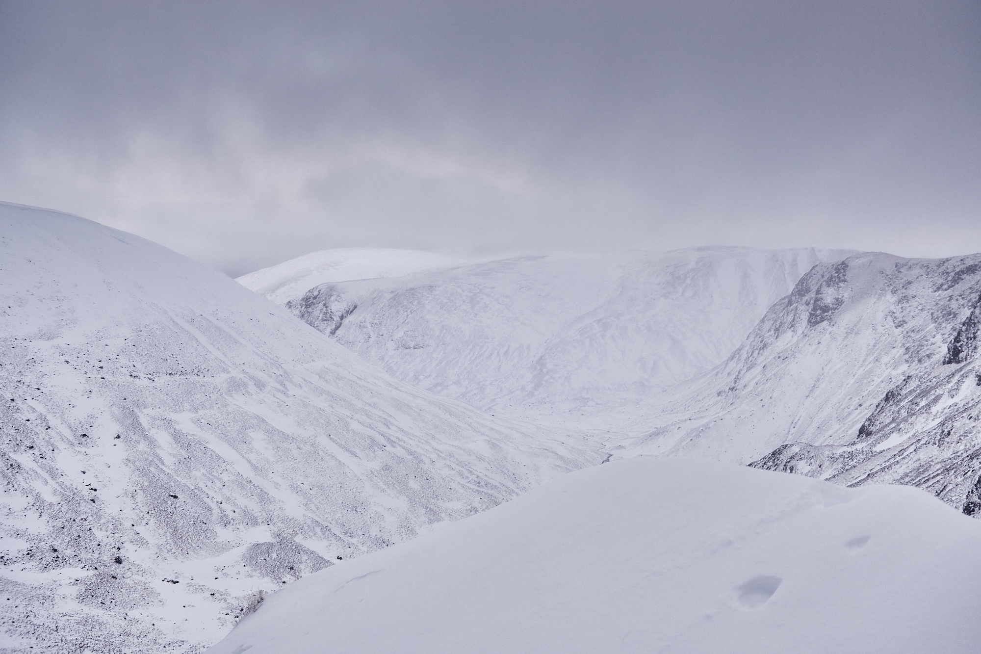 A snowy mountain landscape of rounded hills and dark craggy cliffs with ominous grey clouds above