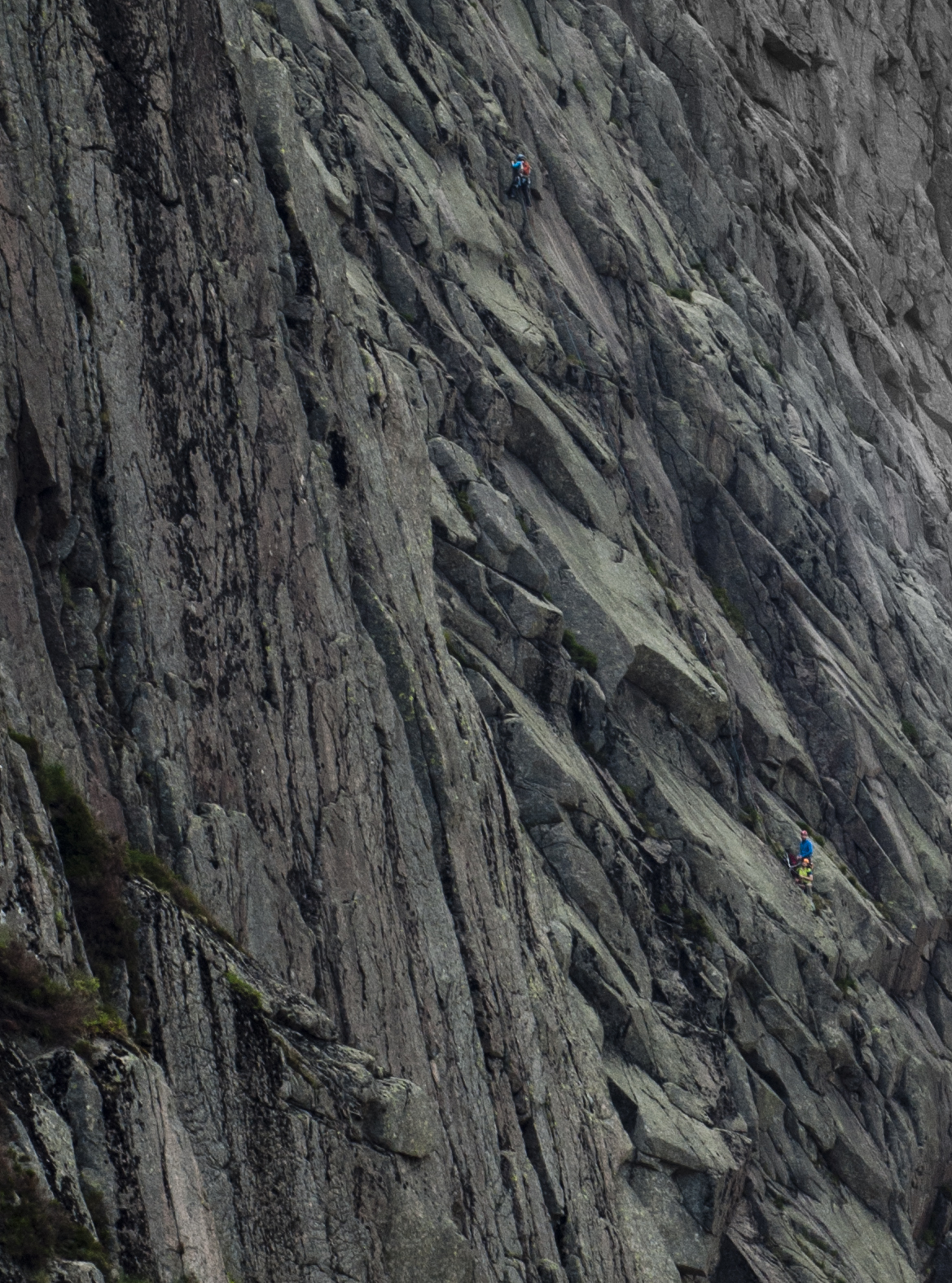 A steep, intimidating, grey and pink granite cliff consisting of slabs and overlaps split by vertical cracks. Rock climbers in brightly coloured clothing are climbing a crack system, joined by a rope