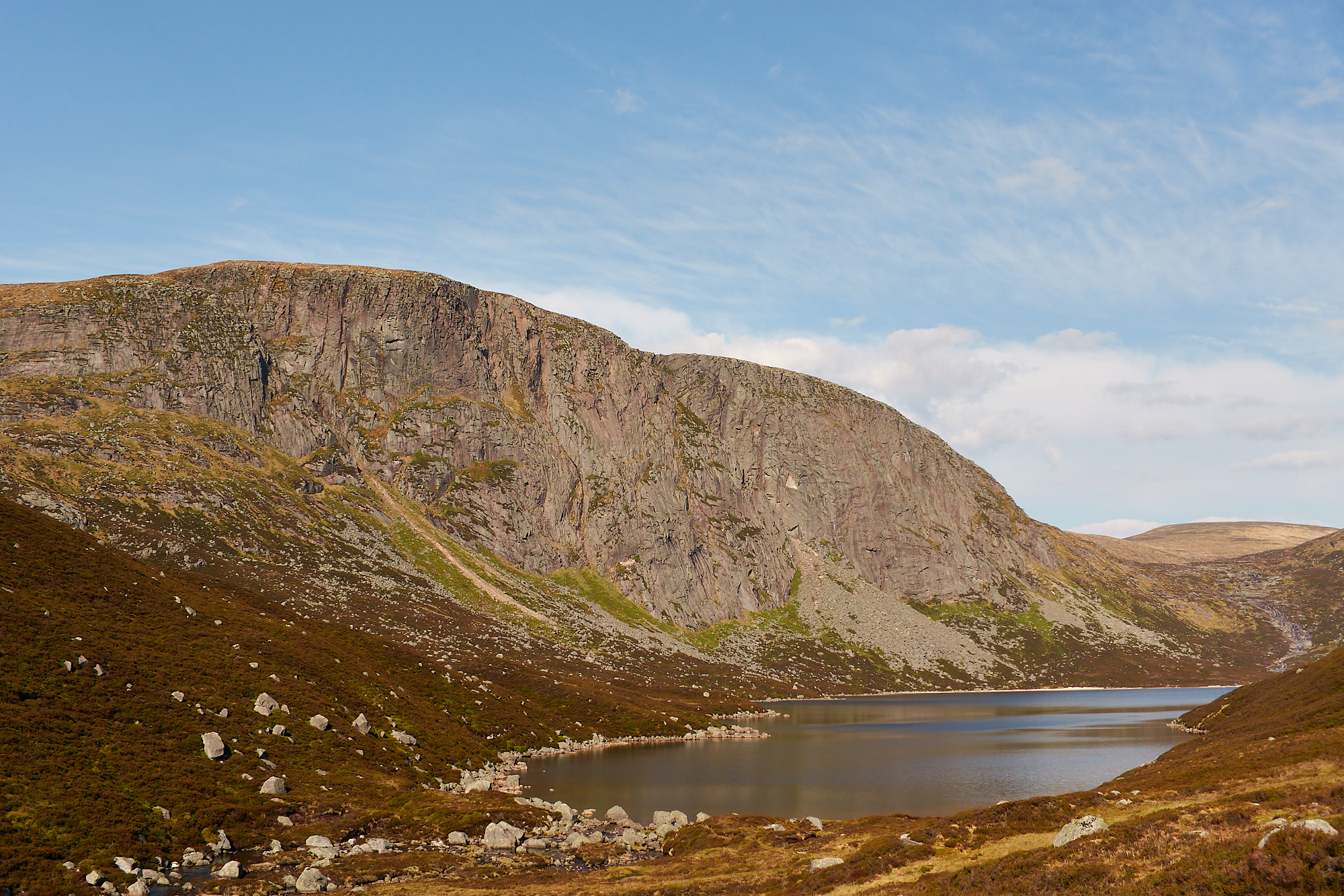 A large pink and grey granite cliff above a Scottish loch surrounded by brown heather beneath a blue sky with scattered clouds