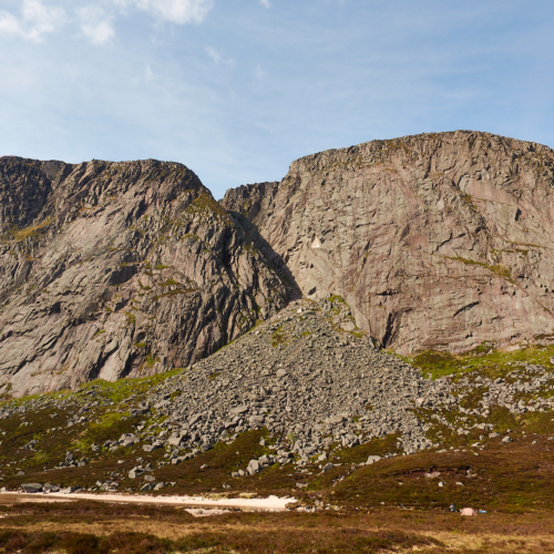 A large pink and grey granite cliff above a Scottish loch surrounded by brown heather beneath a blue sky with scattered clouds. A large boulder field lies beneath a large gully in the middle of the cliff