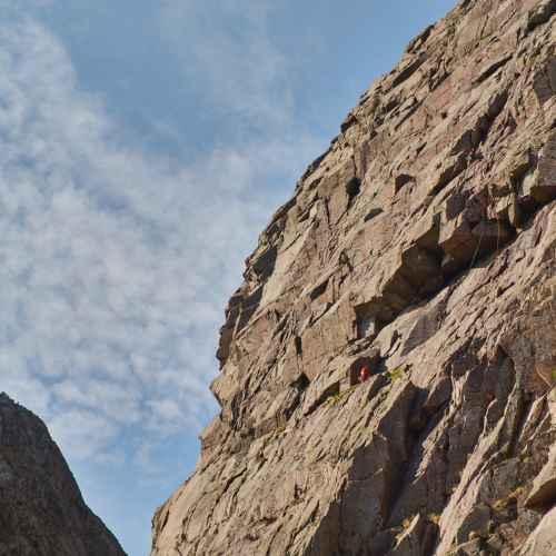 A steep pink and grey granite cliff beneath a blue sky with scattered clouds. Two rock climbers are climbing through a large overhang
