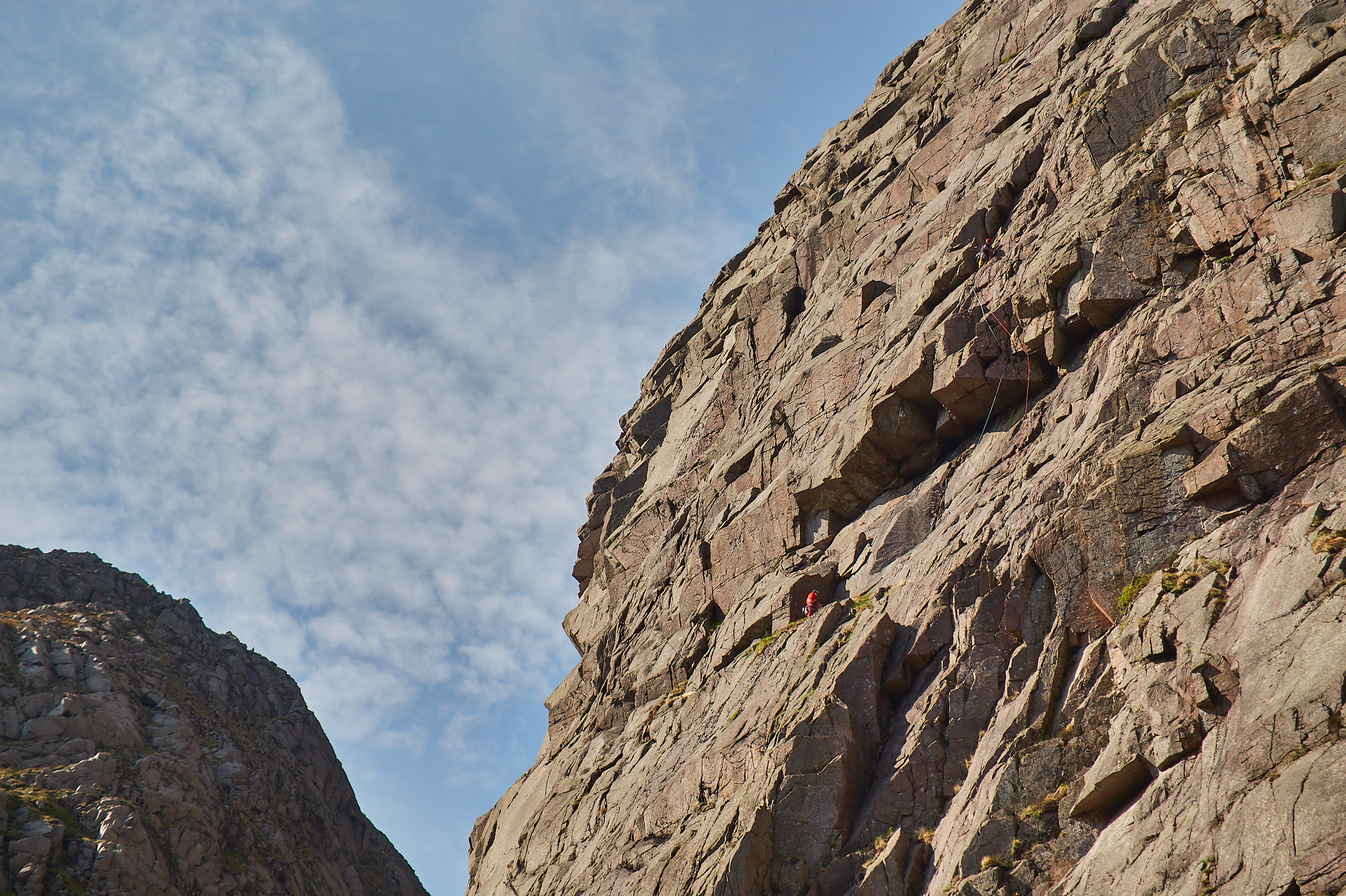 A steep pink and grey granite cliff beneath a blue sky with scattered clouds. Two rock climbers are climbing through a large overhang
