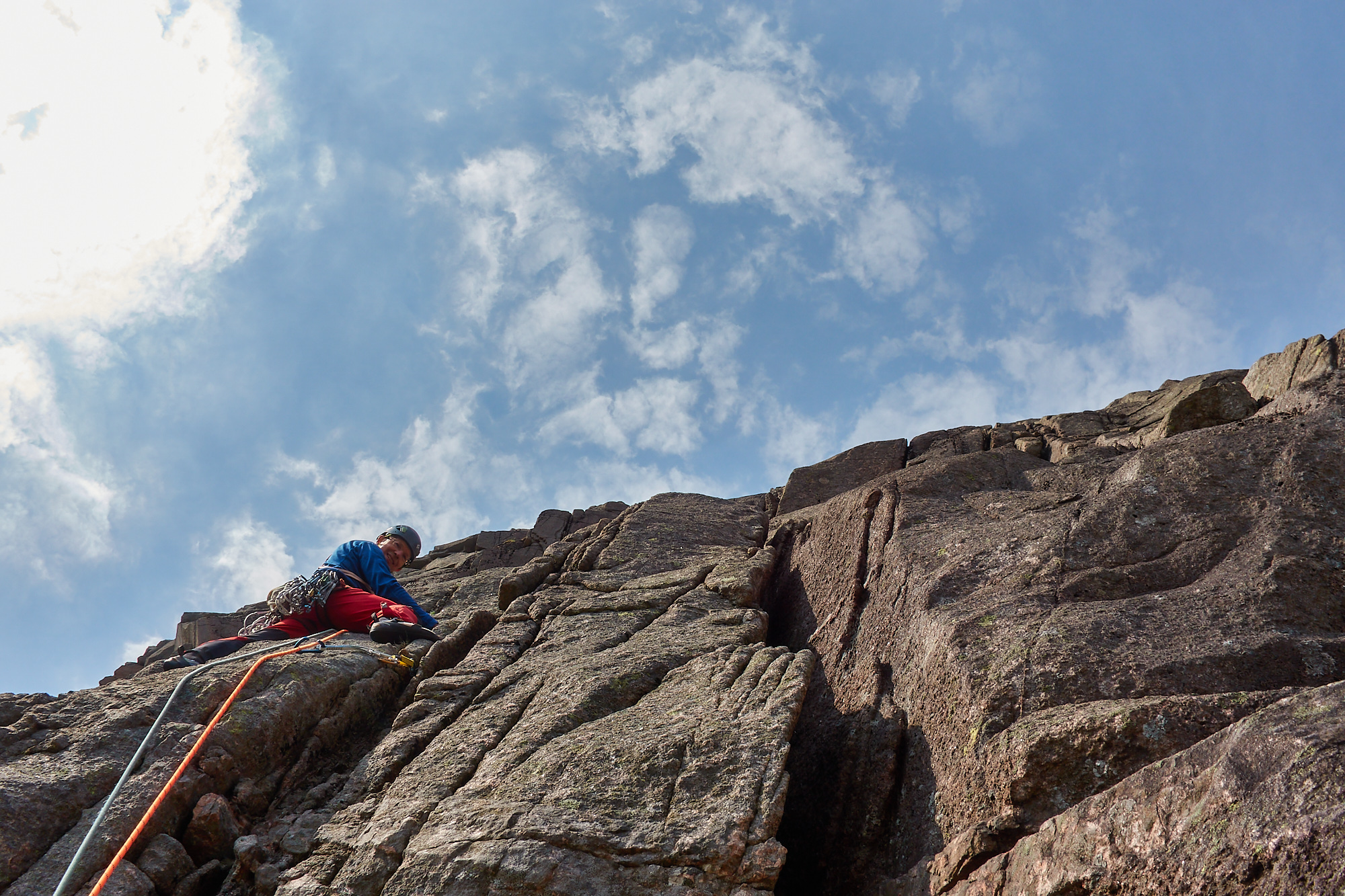 A rock climber in red trousers above a bulge cut by a crack on a steep granite cliff beneath blue sky and scattered white clouds