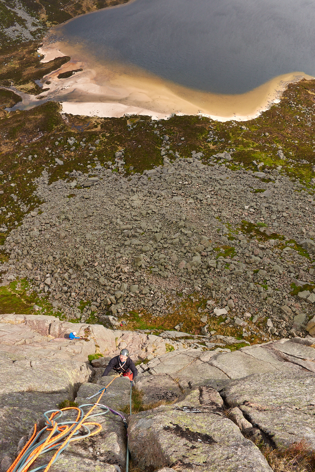Looking down a steep pink granite cliff to a rock climber who is climbing up a crack. A coil of ropes is draped on the rock at the bottom of the photo and a lake with a sandy beach can be seen beneath the cliff