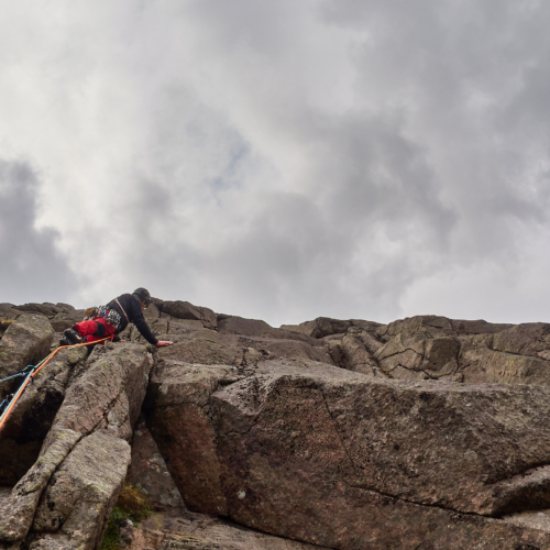 A rock climber in red trousers above a bulge cut by a crack on a steep granite cliff beneath a darkening sky with scattered white and grey clouds