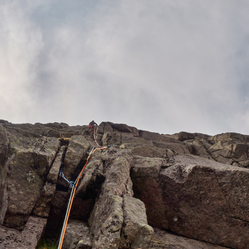 A rock climber in red trousers climbing up rounded cracks on a steep granite cliff beneath a darkening sky with scattered white and grey clouds