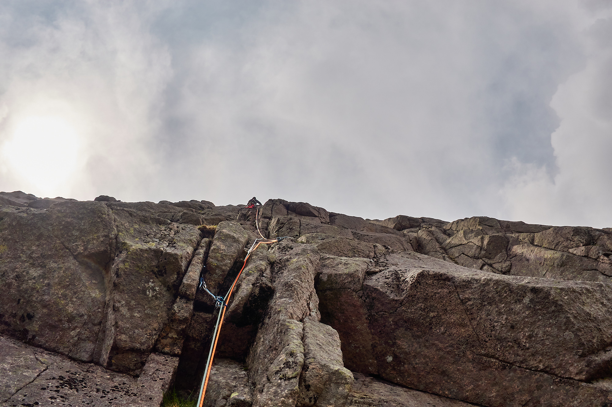A rock climber in red trousers climbing up rounded cracks on a steep granite cliff beneath a darkening sky with scattered white and grey clouds