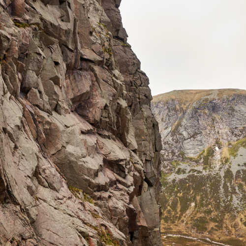 A very steep pink and grey granite cliff with a series of slabs and overlaps cut by vertical cracks. A mountain with more slabby cliffs and heathery slopes lies in the background beneath a grey sky