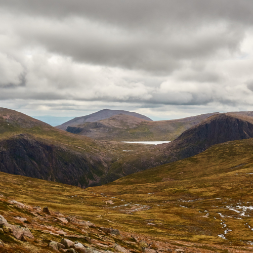 A view of dark craggy cliffs amongst rounded and rugged ancient mountains, covered in green and brown heather and grasses with a moody grey sky above