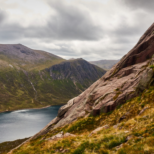 A view of sweeping pale pink granite rock slabs above a dark blue mountain loch, ringed by dark crags and steep mountain slopes beneath a moody cloudy sky
