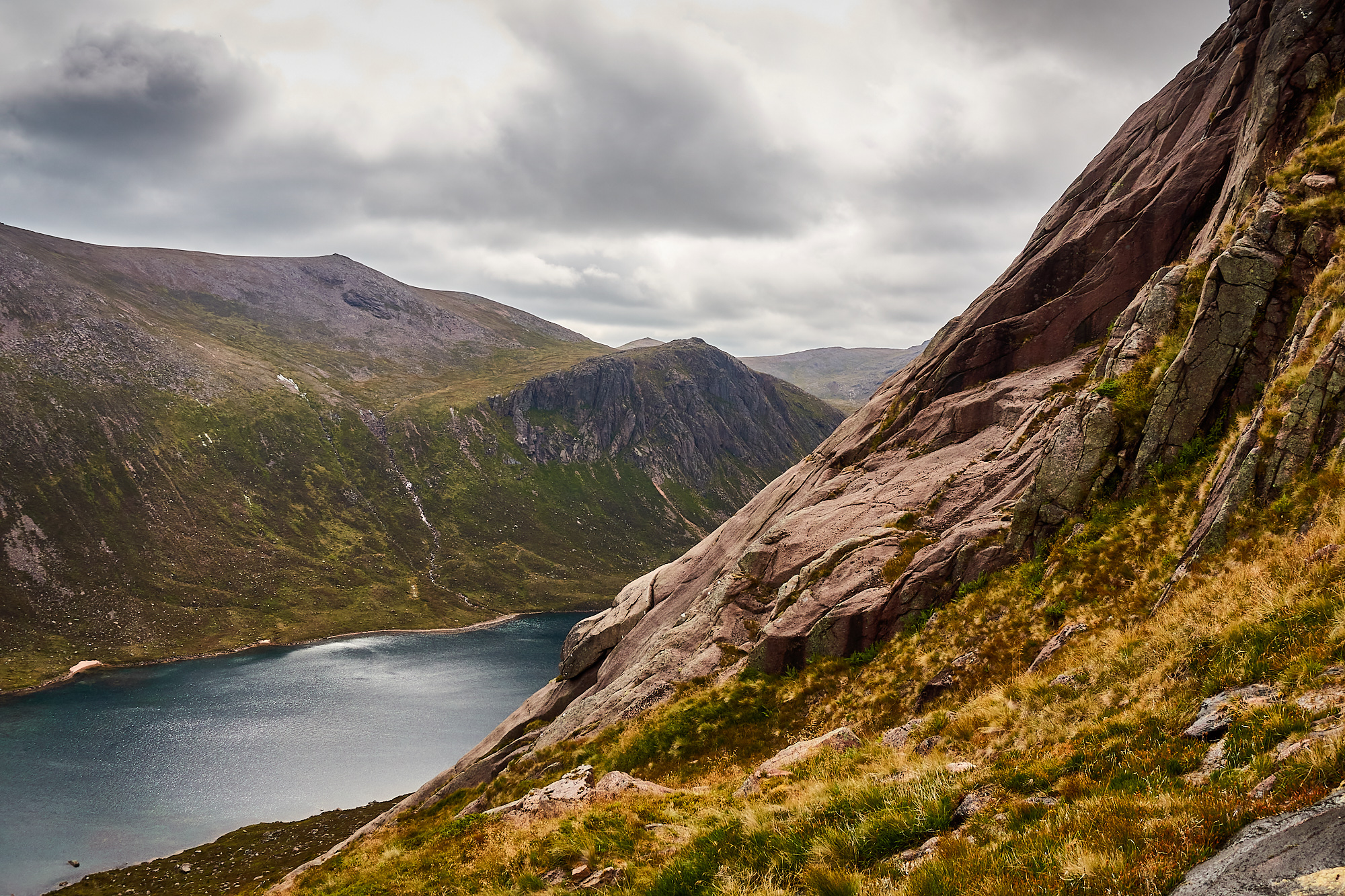 A view of sweeping pale pink granite rock slabs above a dark blue mountain loch, ringed by dark crags and steep mountain slopes beneath a moody cloudy sky