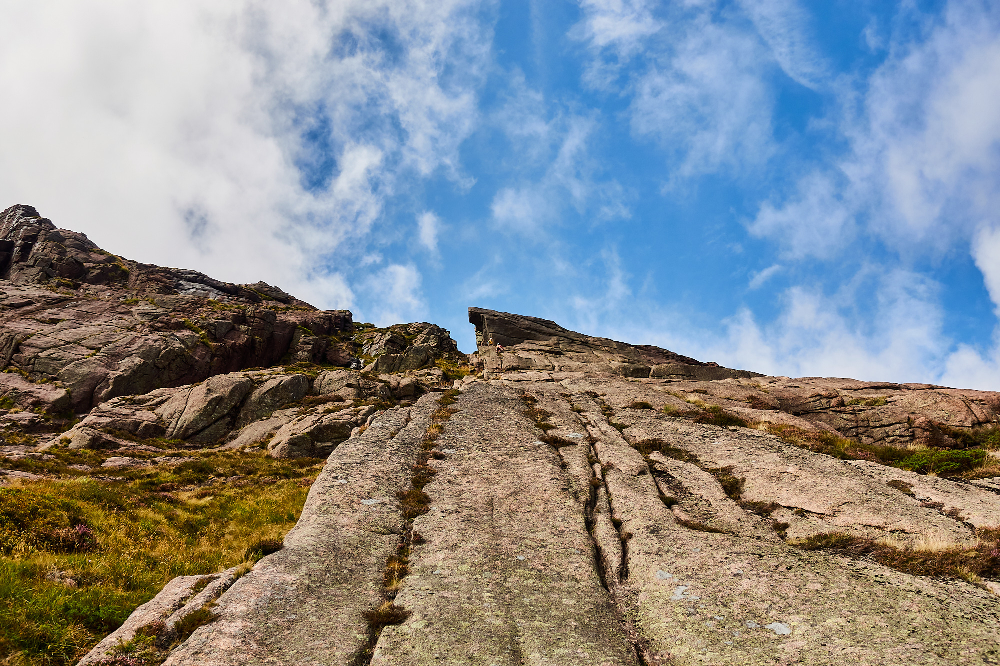 A view of sweeping pale pink granite rock slabs with vertical cracks beneath a cloudy blue sky. Small rock climbers can just be seen at the top of the cliff.