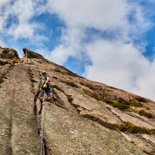 A view of sweeping pale pink granite rock slabs with vertical cracks beneath a cloudy blue sky. A rock climber is just starting to climb one of the cracks and looks back towards the camera with a smile.