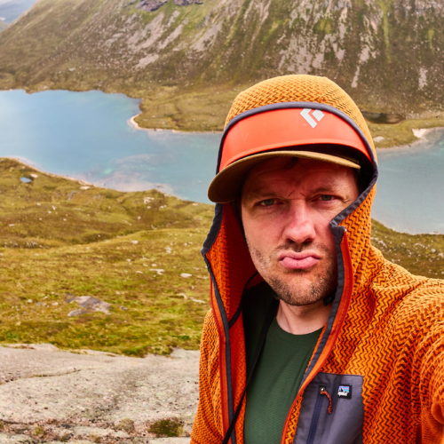 A climber dressed in an orange fleece with a red helmet stands pouting at the camera as he takes a picture of himself, with a blue loch behind and green heather-clad slopes